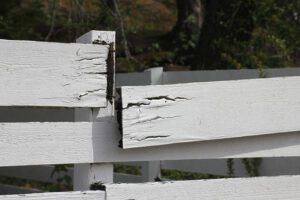 Weathered white wooden fence with peeling paint and cracked boards, outdoors with trees in the background.